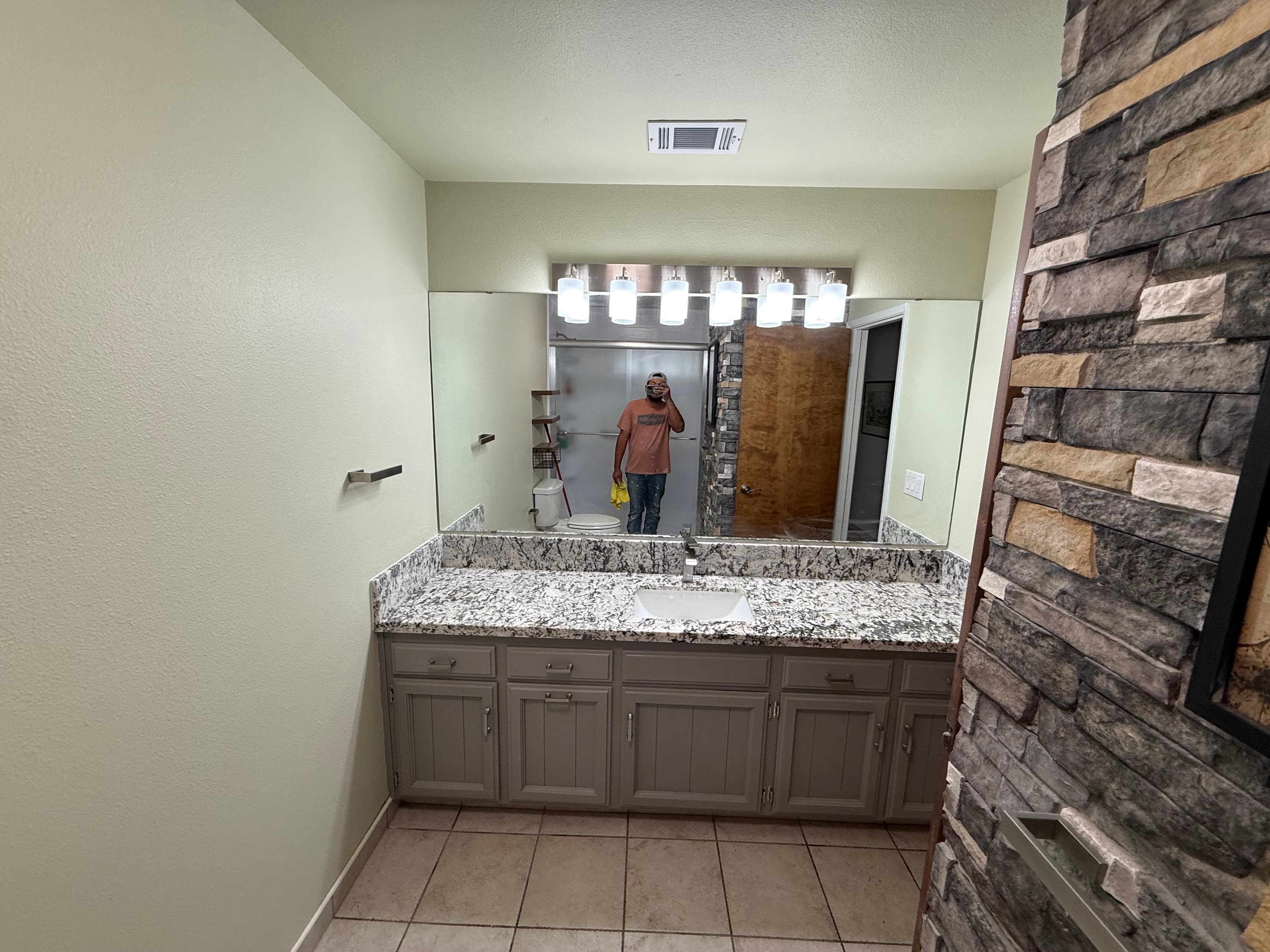 Modern bathroom vanity with gray cabinets, granite countertop, large mirror, and stone accent wall.