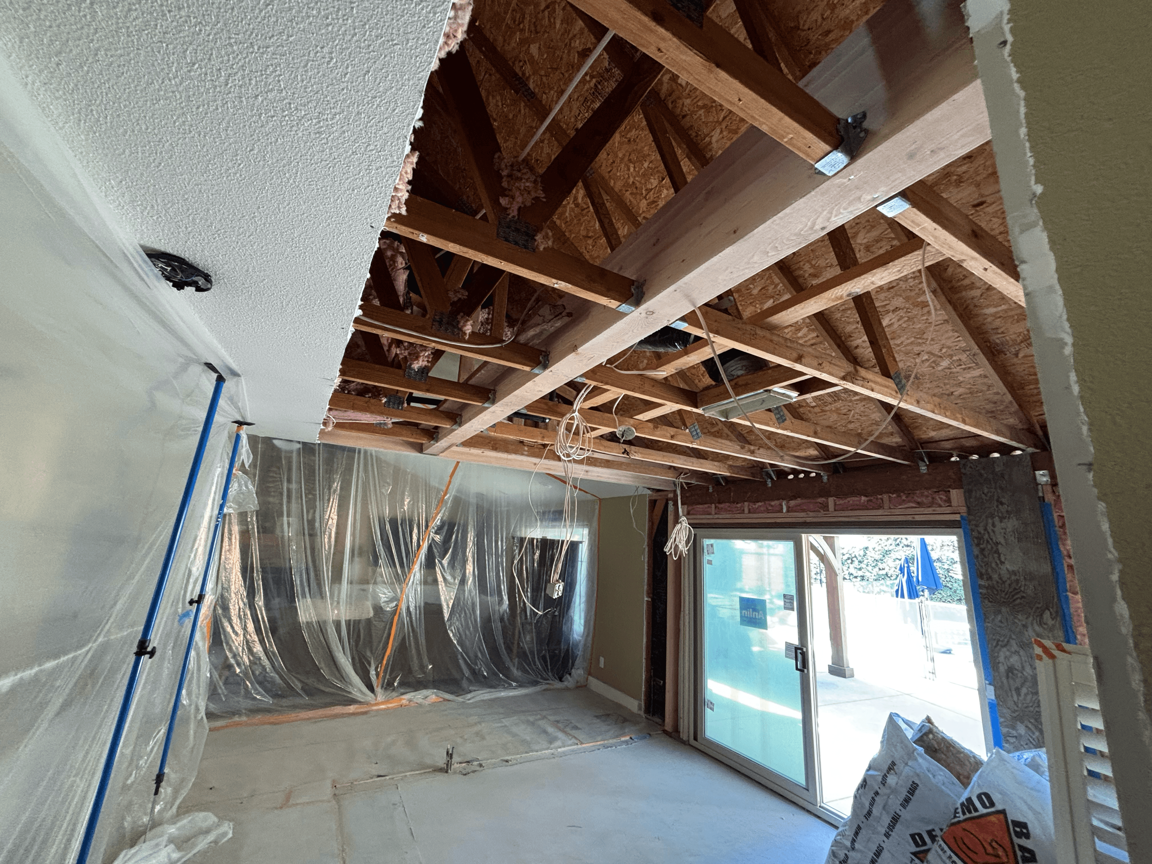 Interior home renovation featuring exposed wooden ceiling trusses, support beams, and plastic protective sheeting.
