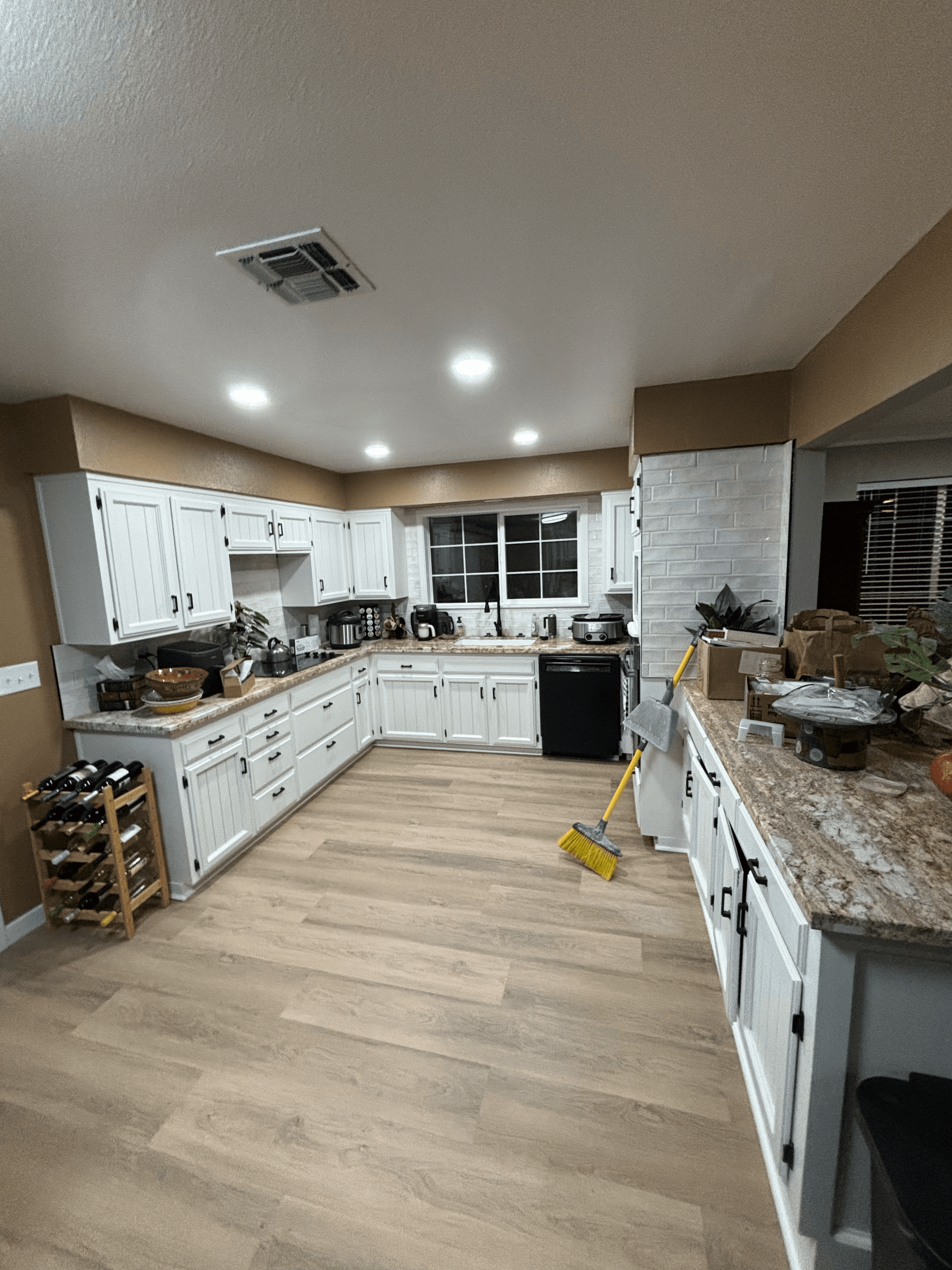 Spacious kitchen with white cabinets, granite countertops, light wood floors, and a yellow broom.