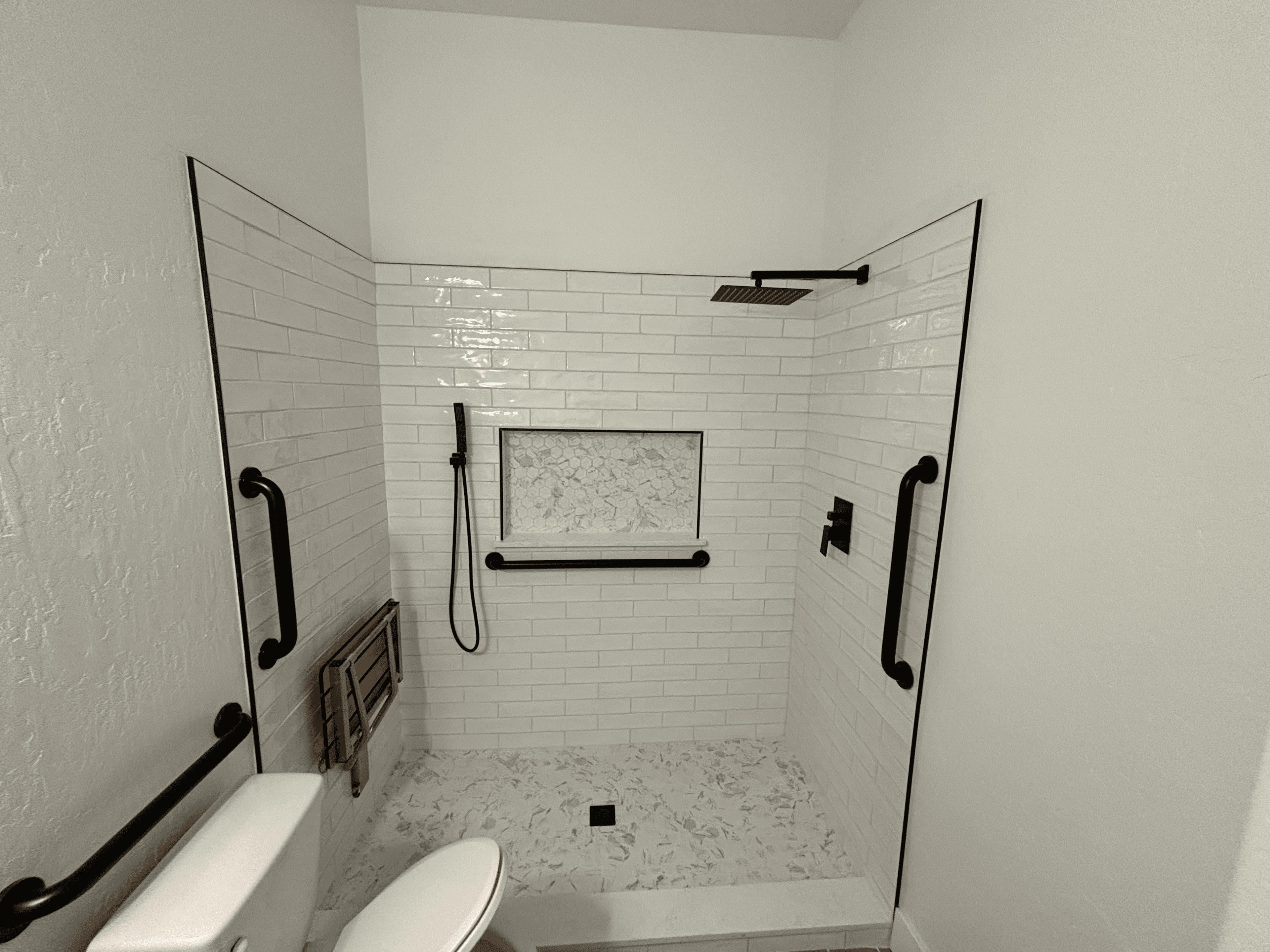 Modern walk-in shower with white subway tiles, black fixtures, marble floor, and folding wooden seat.