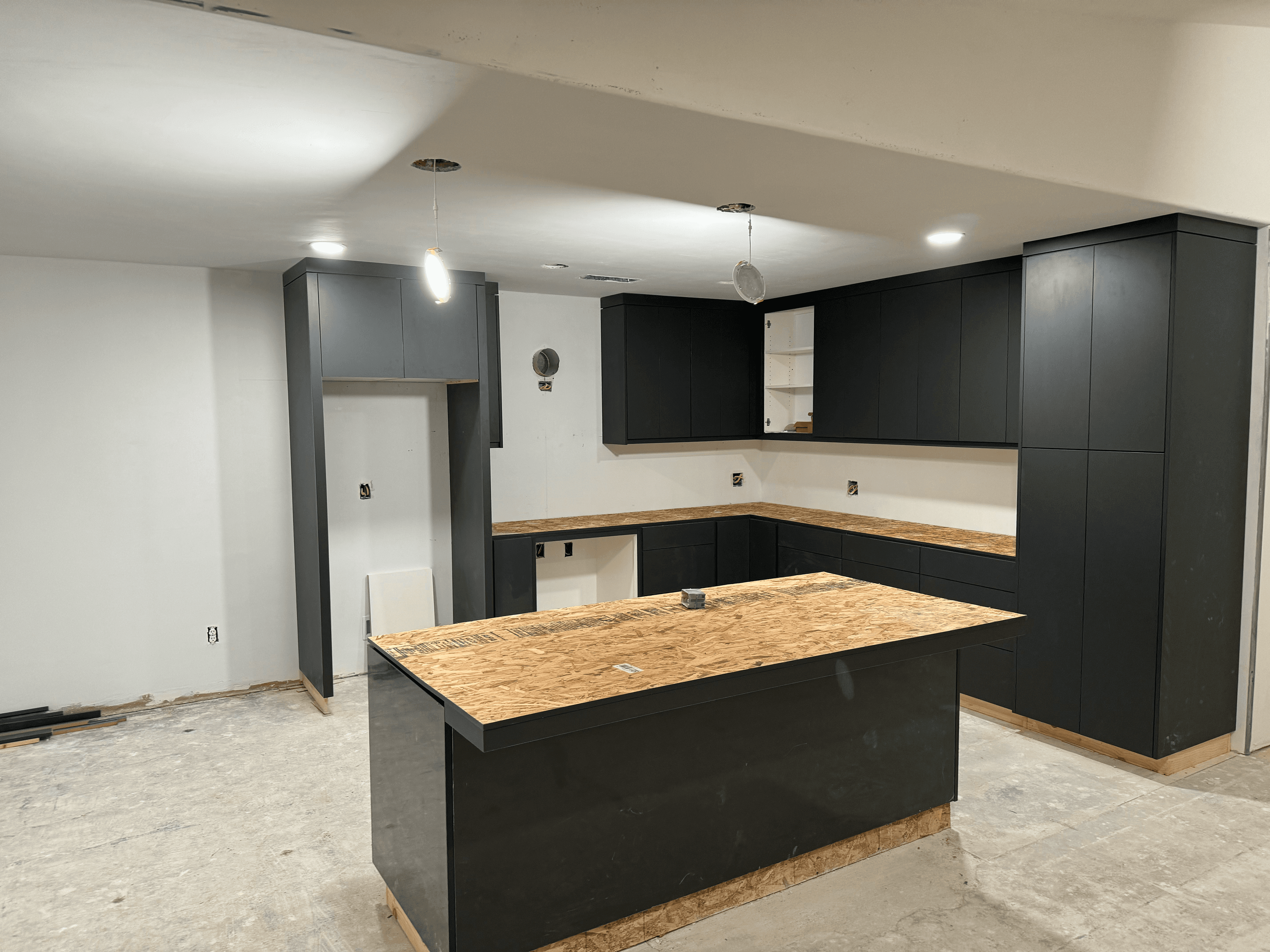 Modern black kitchen cabinets and island with unfinished plywood countertops in a room under construction.